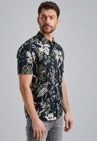 Camisa de manga corta con fondo azul marino oscuro, grandes patrones florales en verde y blanco, y un diseño de cuello clásico. Material de algodón.