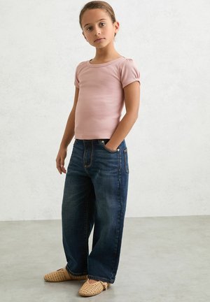 Top roz deschis cu mâneci scurte și model reiat, blugi evazați de culoare albastru închis și pantofi slip-on bej țesuti pe un fundal gri deschis.