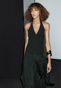 Schwarzes Halter-Tops mit Knöpfen, kombiniert mit einem fließenden schwarzen Rock aus strukturiertem Stoff. Schwarzes Armbandakzent am Handgelenk.