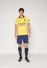 Mann som står i gul AC Milan fotballdrakt, blå shorts, svarte knestrømper og svarte fotballsko mot en enkel grå bakgrunn.
