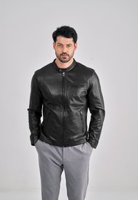 Černá kožená bunda s kulatým límcem, zavíráním na přední zip a bočními kapsami, nošená přes světle šedé kalhoty. Hladká textura a přiléhavý střih.