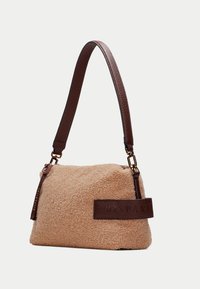 Sac en velours beige avec une anse en cuir marron. Présente un accent en cuir avec un lettrage embossé et des détails en or. Forme rectangulaire.