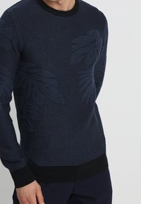 Marinblå stickad tröja med ett texturerat mönster med bladmotiv, rund halsringning samt kontrasterande svarta ribbade muddar och fåll.