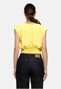 Top giallo a maniche corte con vita elastica. Indossato con jeans in denim scuro dotati di una toppa in pelle e tasche posteriori. Tessuto liscio, vestibilità rilassata.