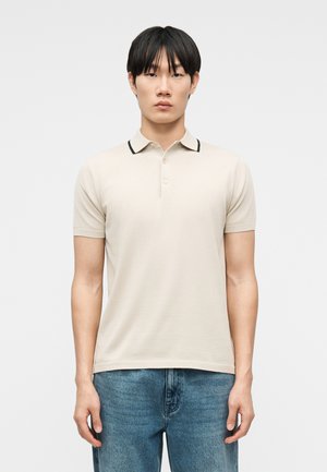 Νέος άντρας με ίσια μαύρα μαλλιά, ντυμένος με μπεζ πόλο μπλουζάκι κοντό μανίκι με μαύρη ρίγα στον γιακά και μπλε τζιν παντελόνι, που στέκεται μπροστά σε απλό φόντο.