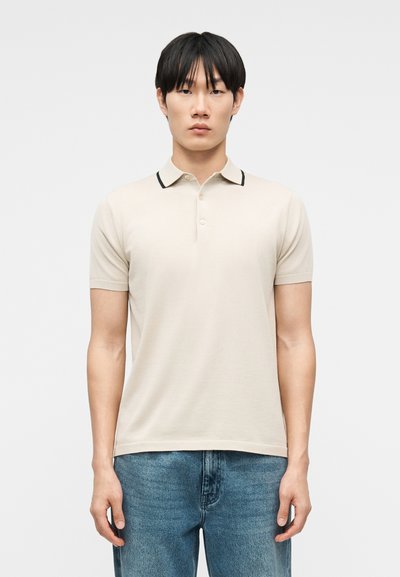 Young man with straight black hair wearing a beige short-sleeve polo shirt with black collar trim and blue jeans, standing against a plain background.