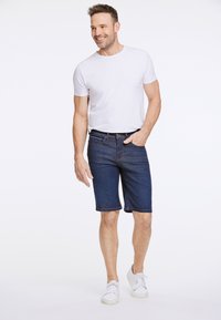 Shorts en denim bleu foncé avec de petites poches, une coupe décontractée et un ourlet retourné. Associés à un t-shirt blanc à manches courtes et des baskets blanches.