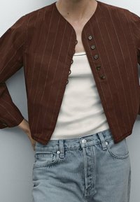 Brun stribet jakke med rund hals, syv knapper og lange ærmer, parret med en cremet top og lyseblå jeans.