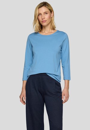 Blonde Frau, die ein hellblaues Langarmshirt und eine dunkelblaue Nadelstreifenhose trägt, steht vor einem einfarbigen hellgrauen Hintergrund.