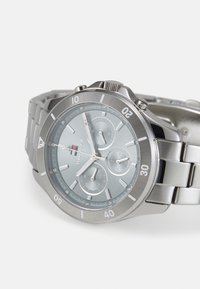 Orologio in metallo argenteo con quadrante blu, tre sottodial e indicatori luminosi. Presenta un bracciale in acciaio inossidabile e una lunetta testurizzata.