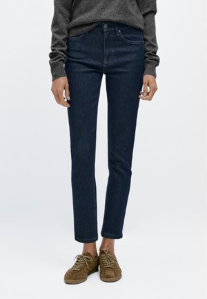 Dark blue denim jeans with a slim fit, mid-rise waist, and classic five-pocket design; paired with brown suede sneakers.