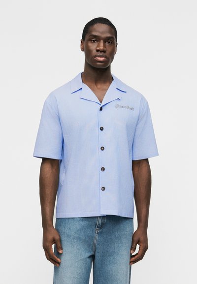 Homme debout face à l'avant, portant une chemise à manches courtes boutonnée bleu clair à fines rayures et un jean bleu clair, sur fond uni.