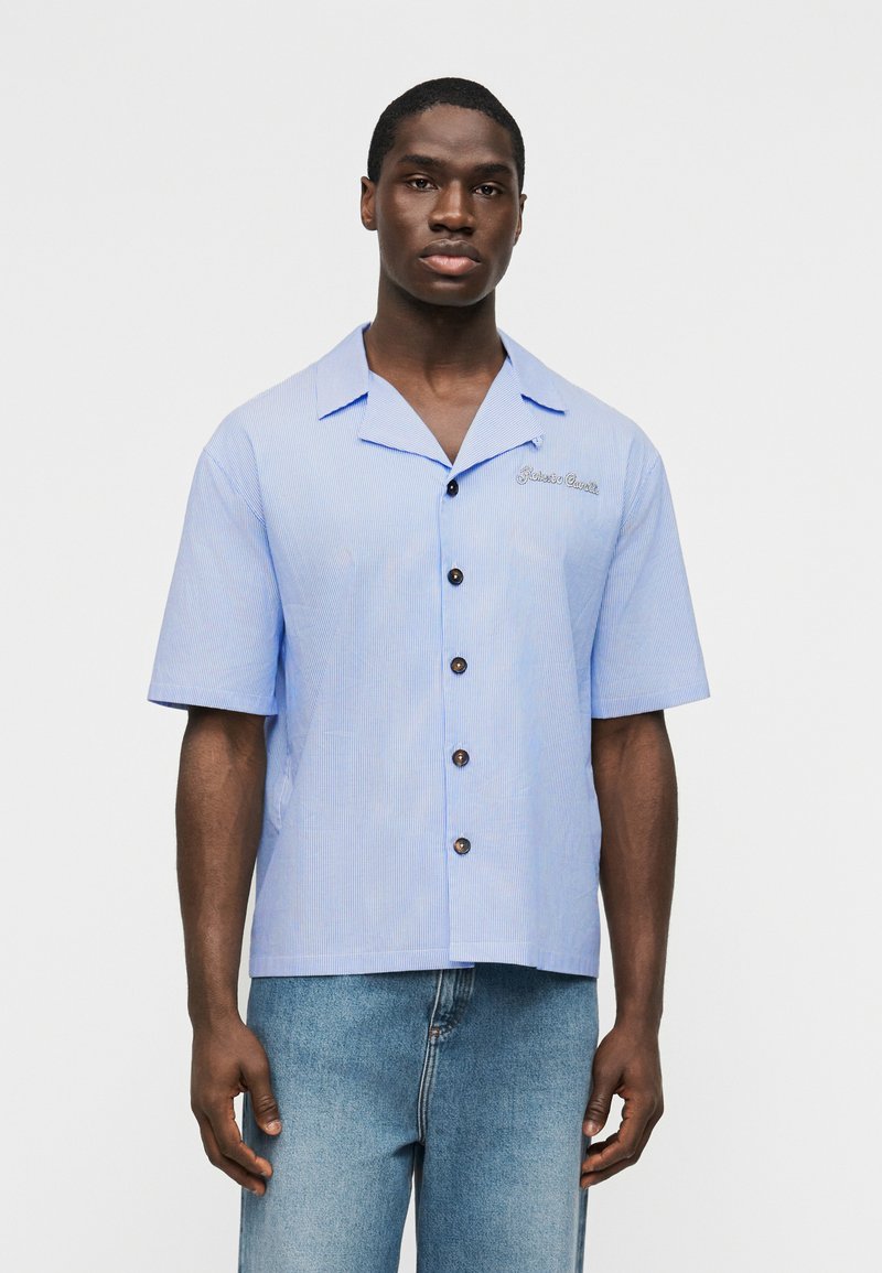 Άνδρας στέκεται με το βλέμμα προς τα εμπρός, φορώντας ανοιχτό μπλε κοντομάνικο πουκάμισο με λεπτές ρίγες και ανοιχτό μπλε τζιν, μπροστά σε μονόχρωμο φόντο.