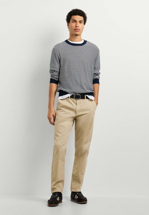 Junger Mann steht mit der Hand in der Tasche, trägt ein gestreiftes Langarmshirt, beige Hose, schwarzen Gürtel und schwarze Schuhe vor schlichtem Hintergrund.