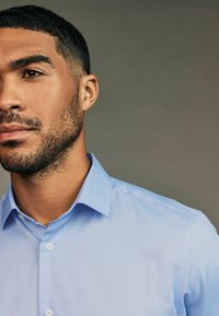 Helles blaues Button-Down-Hemd mit strukturiertem Stoff, Punktkragen und zwei Knöpfen an der Knopfleiste, teilweise an einem männlichen Model gezeigt.