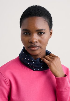 Ung kvinde med kort krøllet hår iført en lyserød sweater, der justerer et mørkeblåt tørklæde med flerfarvede polkaprikkede.