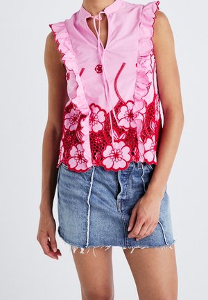 Vrouw draagt een mouwloze roze blouse met rode bloemenborduur en ruches, gecombineerd met een blauwe denim minirok met rafelige zoom.