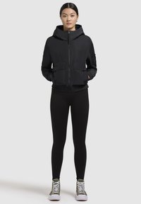 Veste à capuche noire avec fermeture éclair à l'avant, deux poches devant, et poignets élastiques, accompagnée de leggings noirs et de baskets vert olive.