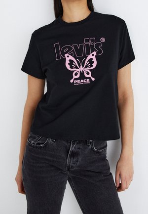Kobieta w czarnej koszulce Levi's z różowym motylem i słowami "Peace comes from within", w połączeniu z ciemnoszarymi dżinsami.