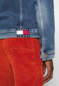 Denimová bunda s modrým efektovým pracím úpravou, sešitá nášivka "TOMMY JEANS". Kombinována s oranžovými manšestrovými kalhotami s žebrovanou texturou.