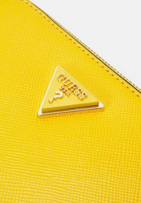 Material giallo testurizzato con un emblema dorato triangolare che presenta la scritta "GUESS USA" in lettere in rilievo e un simbolo di punto interrogativo.