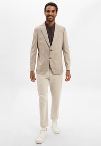 Mann mit Lächeln, bekleidet mit einem beigen Blazer über einer dunklen Zip-Up-Jacke, cremefarbenen Hosen und weißen Sneakers, stehend vor einem schlichten weißen Hintergrund.