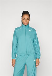 Tyrkysová športová bunda s vysokým golierom, elastickým pásom a bielymi pruhmi. Obsahuje logo Adidas na hrudi.