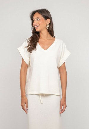 Femme souriante, portant un haut blanc à manches courtes avec col en V et revers retroussés, assorti d'une jupe, sur un fond clair uni.