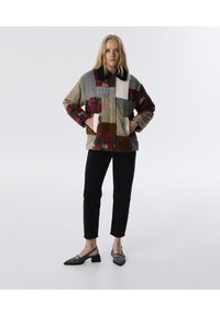 Kleurrijke patchworkjas van verschillende gemusterde stoffen, met een zwarte kraag, zakken en bruine accenten. Zwarte broek en grijze schoenen worden gedragen.