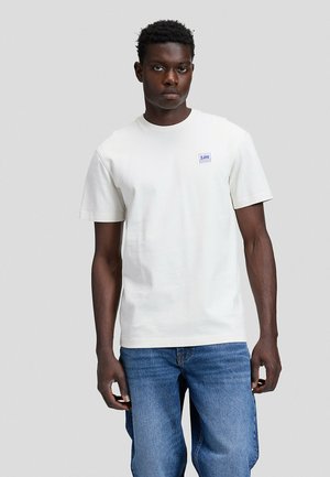 Mann steht frontal, trägt ein schlichtes weißes T-Shirt mit kurzem Ärmel und kleinem Lee-Logo sowie blaue Jeans, neutraler grauer Hintergrund.