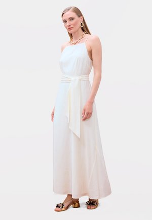 Mujer con un vestido blanco largo sin mangas con cinturón en la cintura, collar de cadena dorada, pendientes de aro dorados y sandalias adornadas con flores doradas.