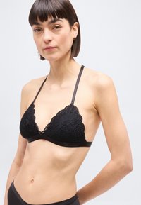 Γυναίκα με κοντά σκούρα μαλλιά που φοράει μαύρο δαντελένιο σουτιέν τύπου bralette και μαύρο εσώρουχο που ταιριάζουν, μπροστά σε απλό φωτεινό φόντο.