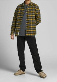 Camicia di flanella a scacchi gialli e neri sopra una maglietta grigia, abbinata a pantaloni neri e scarpe beige. Include chiusura a bottone e vestibilità comoda.