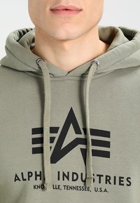 Olivgrüner Hoodie aus Baumwolle, mit Kordelzugkapuze, gerippten Bündchen und großem schwarzem Logo mit geometrischem Design auf der Vorderseite.