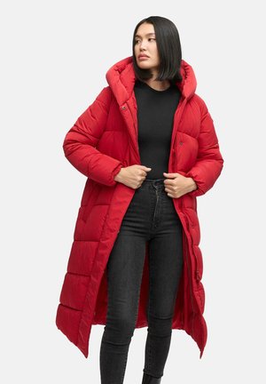 Red long puffer coat with a hood, quilted pattern, and elastic cuffs. Worn over a black top and fitted jeans, featuring a full front zipper.