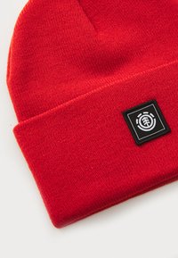 Red knit beanie featuring a folded cuff and a black square patch with circular logo. Soft texture with even stitching throughout.
