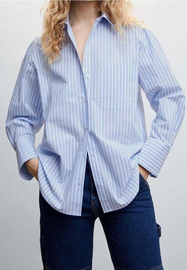 Personne portant une chemise ample à rayures bleu clair et blanc avec des boutons, les mains dans les poches et un jean bleu foncé.