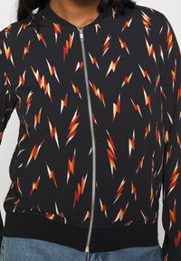 Černá bomber bunda s celoplošným vzorem blesků v červené, oranžové a bílé barvě. Dokončena předním zipem a ribbenými manžetami.
