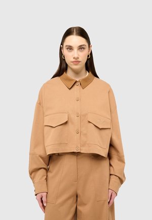 Frau mit langen braunen Haaren trägt eine beige cropped Jacke mit Knöpfen und Klapptaschen sowie passende hoch taillierte Hosen vor grauem Hintergrund.