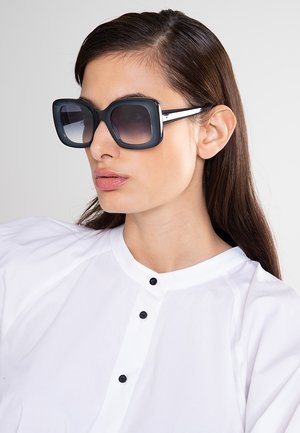 Giovane donna con lunghi capelli castani che indossa grandi occhiali da sole neri quadrati e una camicia bianca abbottonata, guarda di lato.