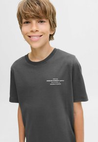 Smiling boy with light brown hair wearing a dark gray t-shirt featuring white text on the chest, against a plain white background.