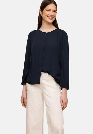 Kvinde smilende iført en langærmet navy blå knapbluse og hvide bukser med højtaljet bred ben, stående foran en ensfarvet baggrund.