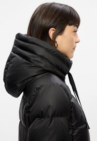Giacca a vento nera con cappuccio alto e ripiegato. Presenta sezioni trapuntate, una texture liscia e un dettaglio con cordoncino. Vista laterale mostrata.