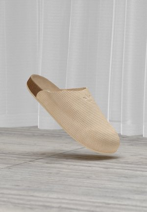 Zapato beige texturizado tipo slip-on con puntera cerrada y parte trasera abierta, situado sobre suelo de madera gris y un fondo de cortina blanca traslúcida.