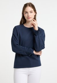 Felpa blu navy con maniche arricciate, tessuto a trama, collo rotondo e polsi aderenti. Abbinata a pantaloni bianchi per un contrasto.