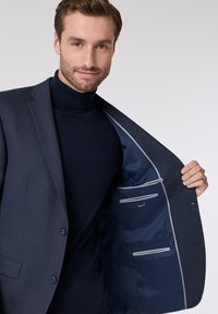 Marineblazer aus strukturiertem Stoff, mit einem einzelnen Knopfverschluss und zwei Innentaschen mit grauen Akzenten. Kombiniert mit einem dunklen Rollkragenpullover.