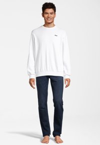 Felpa bianca con logo Fila, scollo rotondo e maniche lunghe, abbinata a jeans blu scuro, taglio aderente e dritto. Nessuna scarpa indossata.