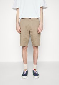 Shorts en coton beige avec une coupe décontractée, dotés d'une fermeture à bouton et de poches latérales. Associés à des baskets navy et vertes, ainsi qu'à des chaussettes légères.