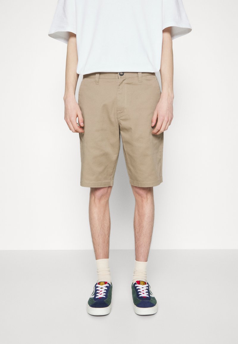 Shorts en coton beige avec une coupe décontractée, dotés d'une fermeture à bouton et de poches latérales. Associés à des baskets navy et vertes, ainsi qu'à des chaussettes légères.