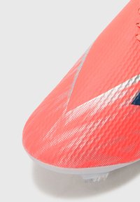 Fényes korall színű futballcipő, textúrált felsőrésszel, ezüst kiegészítőkkel és áramvonalas formával, átlátszó műanyag talpbetéttel.